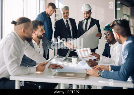 Multiracial group of constructors, builders, engeneers and architects discussing blueprint at office. All men are dressed in business suits, and white Stock Photo