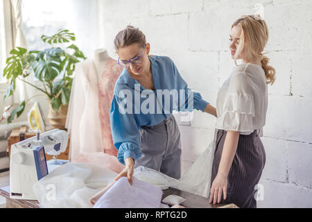 Young beautiful blonde woman chooses fabric for her wedding dress and female professional tailor in stylish blue blouse helps her in Stock Photo