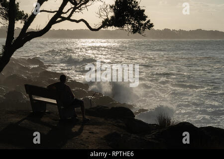 TAURANGA NEW ZEALAND - FEBRUARY 10 2019; Silhouette of man on seat viewing of storm waves crashing into rocky foreshore surrounded by sea spray at bas Stock Photo