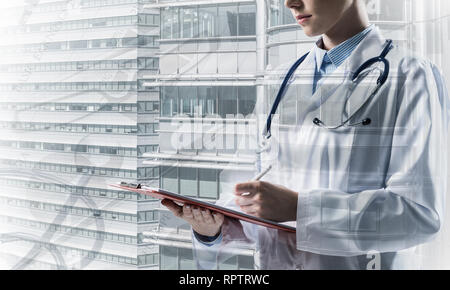 Close up of young woman doctor in white medical suit writing in notebook while standing against city view and medical stuff on background. Double expo Stock Photo