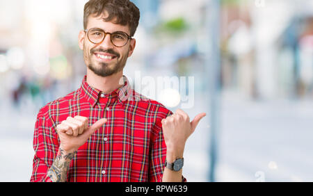 Young handsome man wearing glasses over isolated background Pointing to the back behind with hand and thumbs up, smiling confident Stock Photo