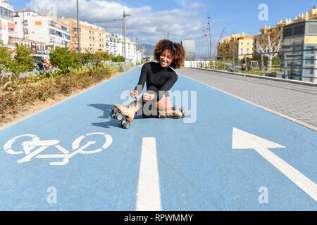 Young smiling black girl sitting on bike line and puts on skates. Stock Photo