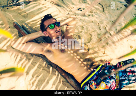 Vacation concept. Happy young adult man swinging at palm grove. Stock Photo