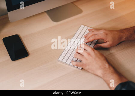 Top view close up of man's fingers typing on a wireless keyboard, set on a wooden working desk. Stock Photo