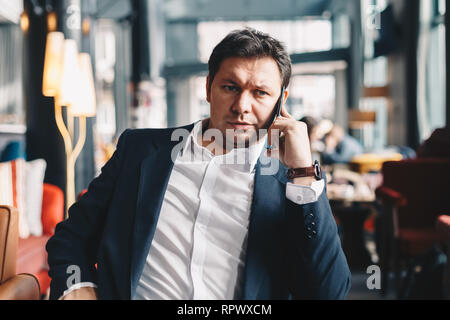 Young good looking male manager sitting in a restaurant while having a business call with his partner, holding a smartphone. Concept of working on a b Stock Photo
