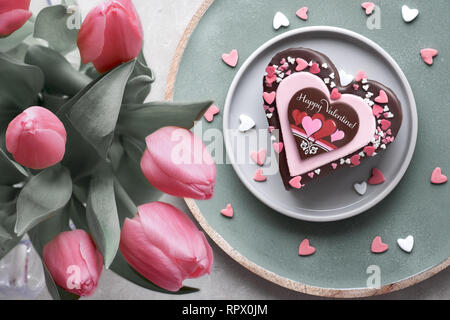 Valentine heart cake with chocolate, sugar decorations and text 'Happy Valeitine' and a bumch of pink tulips. Top view on light background. Stock Photo