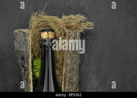 Abstract composition of closed with beige cork champagne black glass bottle placed in tree bark with bent and moss on black stone background Stock Photo