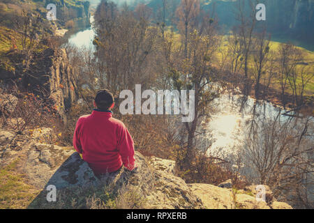 A man sits on a rock above a beautiful canyon on a sunny autumn day. The man looking at the river Stock Photo