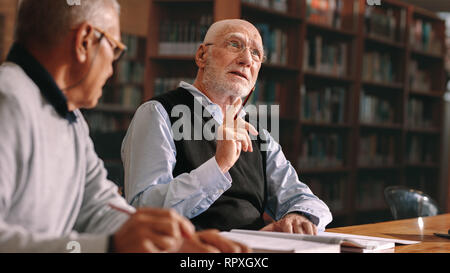 Senior classmates sitting in library and talking. Senior man learning sitting in classroom with his colleague. Stock Photo