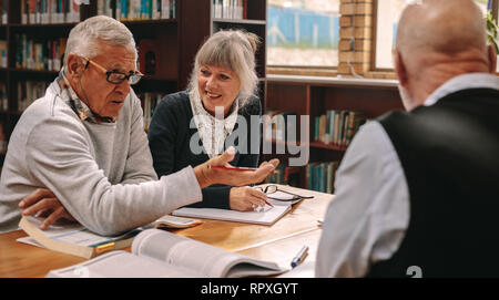 Two senior men and a woman sitting in a library with course books on table and discussing. Senior men and a woman sharing ideas and discussing subject Stock Photo