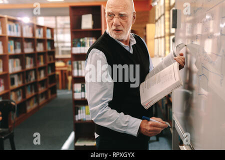 Senior lecturer teaching in class writing on whiteboard holding a book in hand. Professor looking back while writing on board standing in classroom. Stock Photo