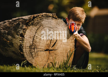 Preteen boy with toy gun prepare for attack and play outdoors. Young child with toy gun hiding behind a log outdoors in park.