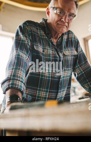 Senior man with protective eye wear standing by workbench and looking at camera. Mature carpenter at his carpentry workshop. Stock Photo