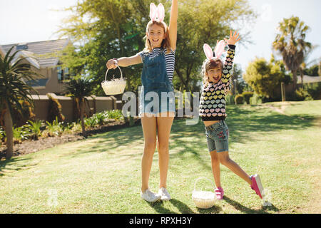 Two kids playing in a garden holding baskets. Girls wearing fancy rabbit ear headband and playing outdoors on a sunny day. Stock Photo