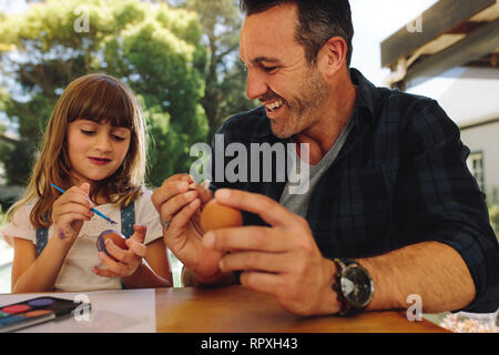 Girl painting an easter egg using a paint brush sitting with her father. Man helping his daughter in painting eggs. Stock Photo