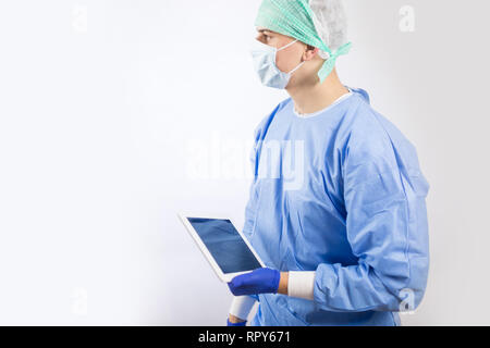 Surgeon doctor in sterile gloves preparing for operation using tablet computer. He is wearing surgical cap and blue gown Stock Photo