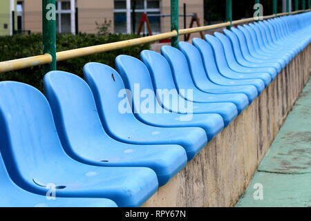 Row of new blue plastic seats mounted on top of concrete bleachers with metal fence and garden vegetation in background next to basketball court Stock Photo