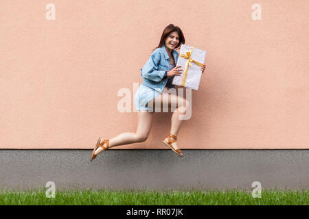 Full length portrait of happy excited beautiful woman in casual jeans denim in summertime standing near sandybrown wall copyspace. holding gift box, j Stock Photo