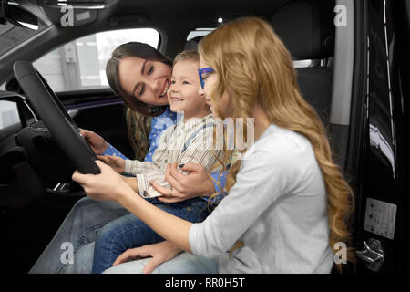 Mother sitting with children in car cabin of new automobile, observing and testing auto. Woman holding cute, happy son on knees, beautiful daughter holding hand on steering wheel. Stock Photo