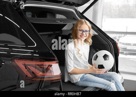 Charming and cheerful girl sitting in car trunk, looking at camera, holding soccer ball, smiling and posing. Beautiful, happy girl wearing glasses, has curly blonde hair. Stock Photo