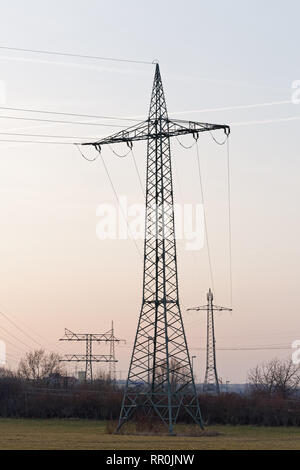Several large electricity pylons in different types with cables, they are staggered as silhouettes in front of a slightly colored evening sky with con Stock Photo