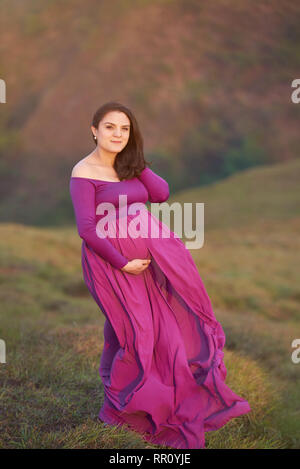 Pregnant woman in pink dress standing on natural sunset background Stock Photo