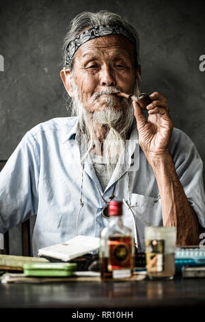 Portrait of an elderly Japanese man smoking a pipe while on the table there is a bottle of rum and painting tools. Osaka, Japan. Stock Photo