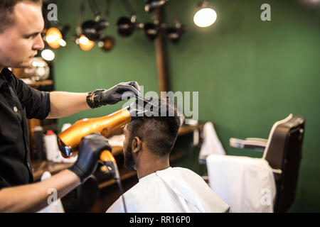 Close up side view of young bearded man getting groomed by hairdresser with hair dryer at barbershop Stock Photo