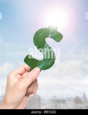 Concept of green gold and green resource, female hand holding a leaf in form of dollar sign shape, on sunlight sky background. Stock Photo