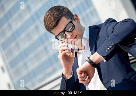 Young man in sunglasses leaning on car talking on smartphone looking at watch time for meeting close-up Stock Photo