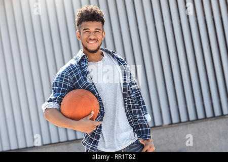 Sports Hobby. Mulatto guy standing isolated on city street with ball smiling playful Stock Photo