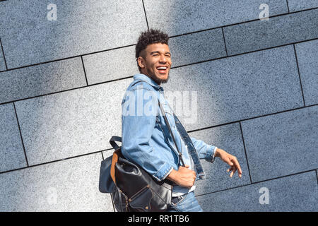 Freestyle. Mulatto guy carrying backpack walking isolated on wall laughing cheerful side view Stock Photo