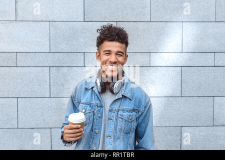 Freestyle. Mulatto guy with headphones on neck standing isolated on wall with cup of coffee smiling toothy Stock Photo