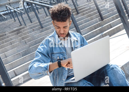Time management. Mulatto guy sitting on stairs using laptop checking time thoughtful cllose-up Stock Photo
