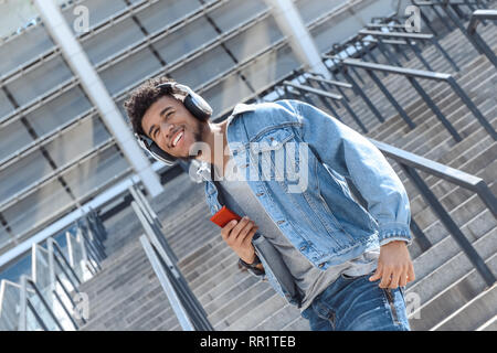 Outdoors Leisure. Mulatto guy in headphones standing on stairs with smartphone listening music smiling happy Stock Photo