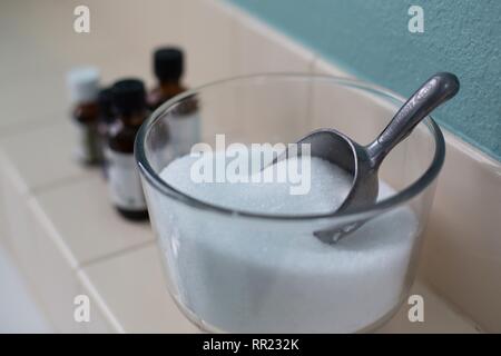 A bowl of epsom salts with a scoop and essential oils,  next to a bathtub. Stock Photo