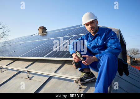 Portrait of male engineer in blue suit and protective helmet installing solar photovoltaic panel system. Electrician looking to the camera, sitting on roof. Alternative energy ecological concept. Stock Photo