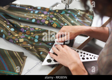 Tailor workplace: close-up of female hands making marking on garment details using chalk and ruler before sewing on electrical sewing machine. Handmade exclusive fashionable clothes creating. Stock Photo