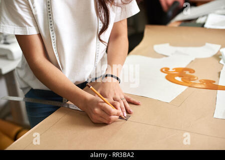 Workplace in design studio, pattern creation process. Female designer hands close-up making drawing lines on cardboard using pencil and ruler. Exclusive clothes making, creative occupation concept. Stock Photo