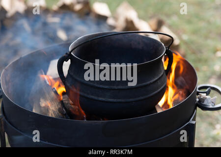 Preparation of food on an open fire. Black metal pot, fire and flames, smoke, stack of wood in the background Stock Photo