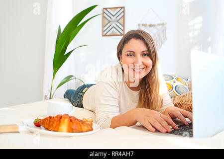 Smiling female freelancer working on laptop and having breakfast in bed at home Stock Photo