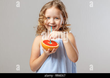 Happy little girl child holds half a grapefruit and drinks juice from a tubule. Healthy food. Gray background, studio Stock Photo