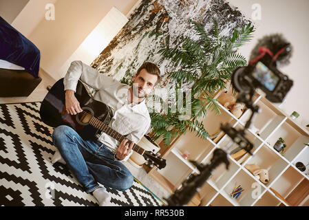 More subscribers. Bearded male music blogger sitting on the floor and holding the guitar, while recording new video for vlog. Youtube. Instagram. Musi Stock Photo