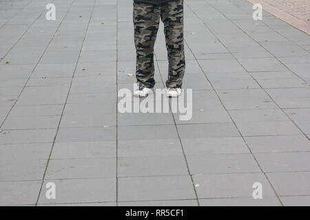 Men feet in old white sneakers and camouflage trousers on pavement on the street. Stock Photo