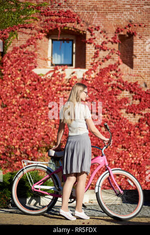 Back view of young pretty long-haired blond woman in casual clothing and glasses posing at pink lady bike on bright warm sunny day on background of brick wall overgrown with beautiful red ivy leaves. Stock Photo