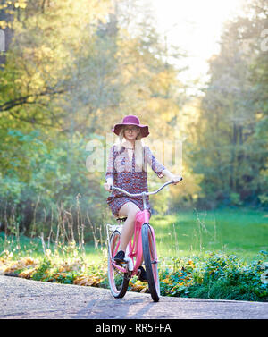 Slim blond fashionable attractive woman in glasses, short dress and pink hat riding lady bicycle along paved park alley on beautiful green and golden trees lit by bright autumn sun background. Stock Photo