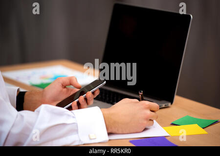 Young manager man using mobile phone in office and makes notes on paper sticker . Close-up hands, table, laptop. Stock Photo