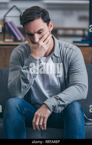depressed man sitting, covering mouth with hand and grieving at home Stock Photo