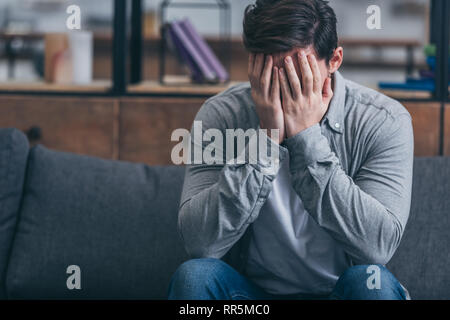 man sitting on couch, crying and and covering face with hands at home Stock Photo