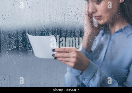 cropped view of grieving woman holding photograph and crying through window with raindrops Stock Photo